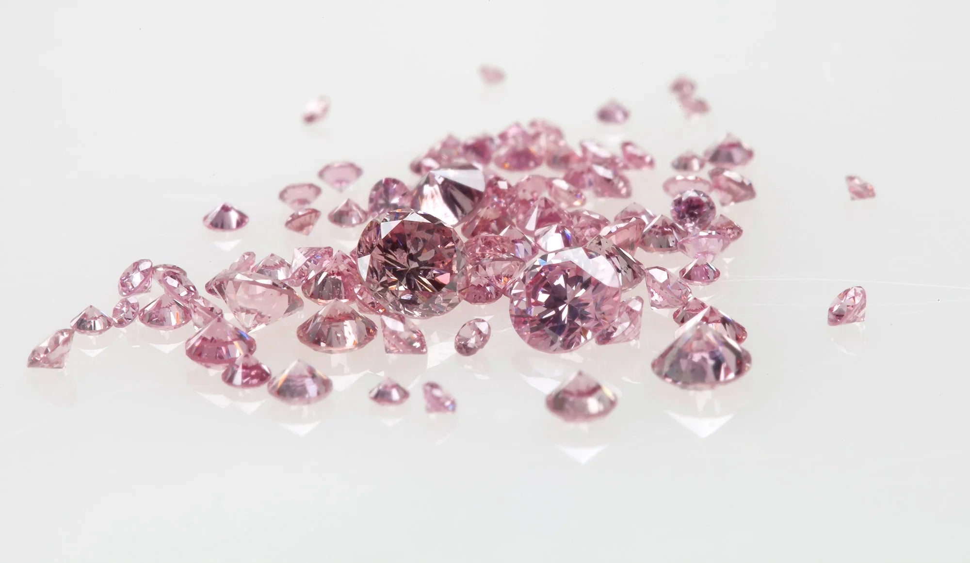 A scattered collection of round brilliant-cut pink diamonds of various sizes, displayed on a reflective white surface, highlighting their vivid pink color and sparkling clarity.